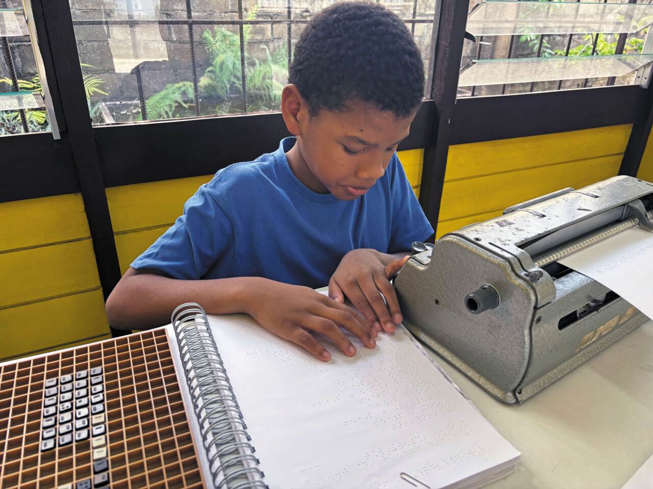 Blinde jongen met brailleer apparaat bij CBB Suriname